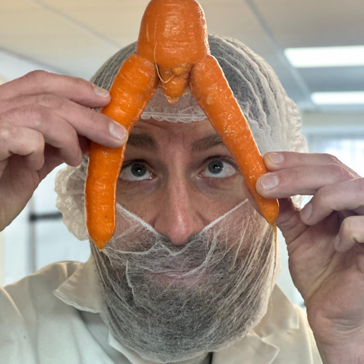 A man wearing a white hairnet and lab coat holds a carved orange carrot shaped like a small figure above his face with both hands in a bright indoor setting.