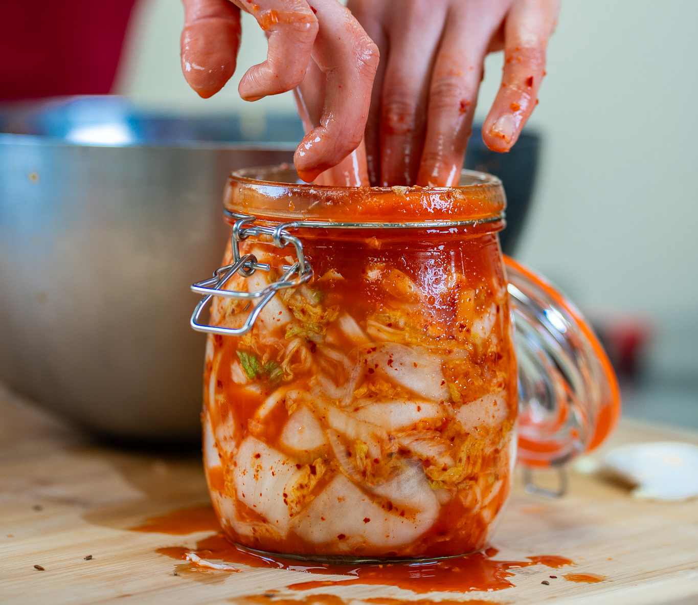 Glass jar of kimchi with a hand reaching in, on a wooden surface.