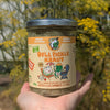 A hand holding a glass jar of Dill Pickle Kraut with cucumber and dill, set against a blurred background of yellow flowers.