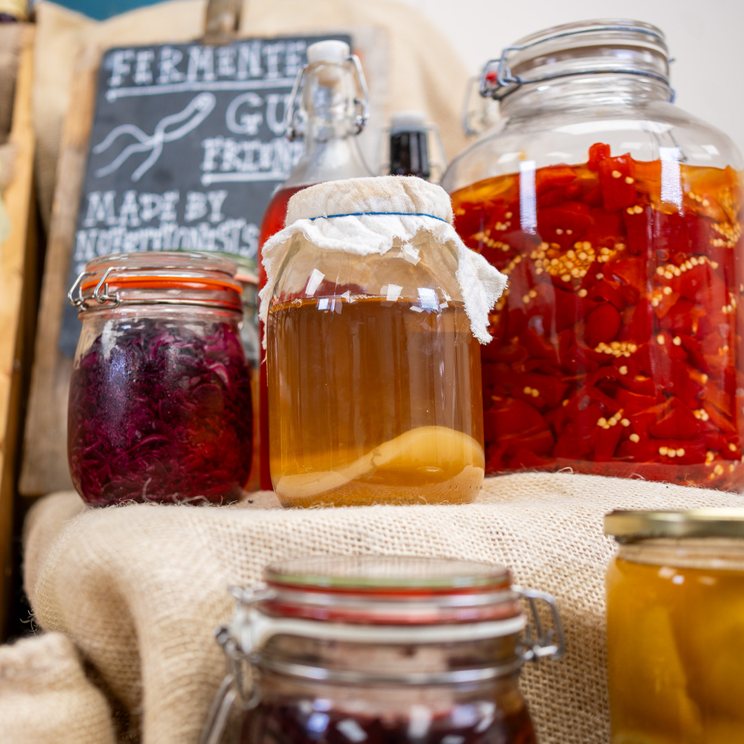 A collection of glass jars filled with various fermented foods and preserves, arranged on a burlap cloth with a chalkboard sign in the background 