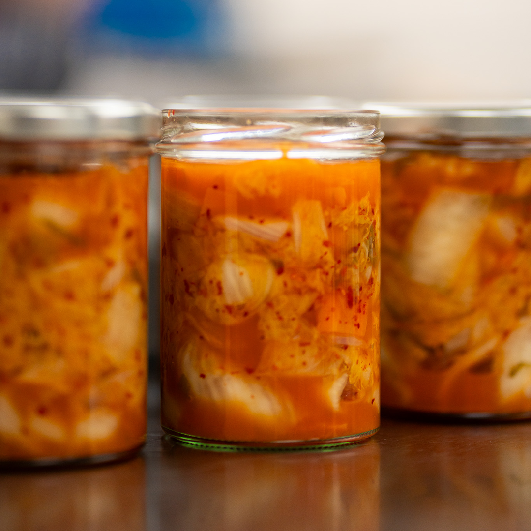 Three jars of kimchi on a reflective surface with a blurred background