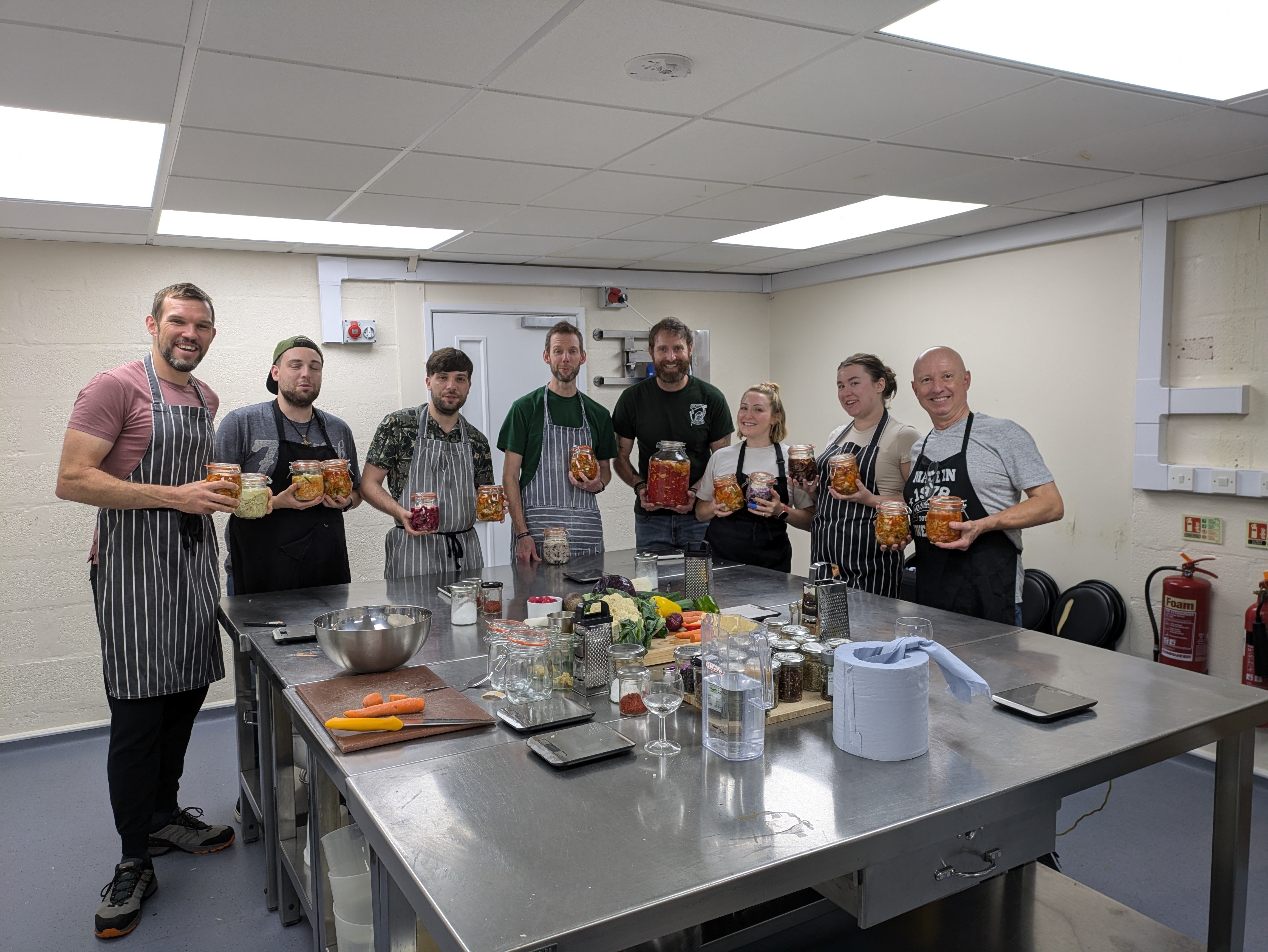 Group of people in a kitchen setting with aprons, standing around a table with food and drinks.